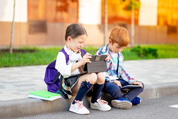 Ein Junge und ein Mädchen mit Aktentaschen oder Rucksäcken sitzen in der Schule und lesen ein Buch und ein Tablet, das zurück in die Schule geht, Bruder und Schwester lernen, Hausaufgaben auf der Straße zu machen