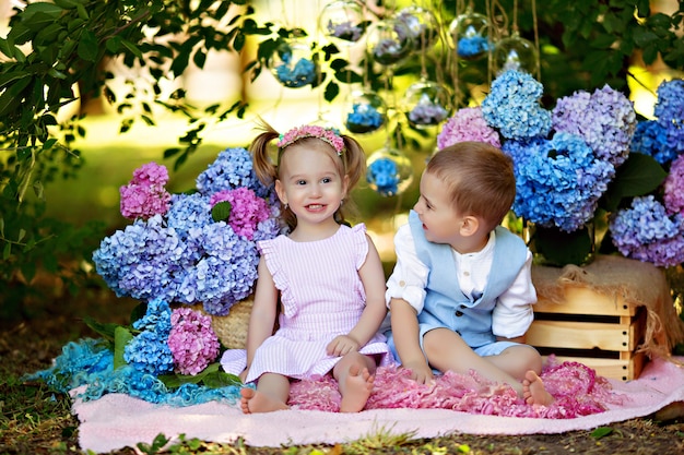 Ein Junge und ein Mädchen sitzen mit Blumensträußen aus Hortensienblumen im Gras der Natur. Bruder und Schwester umarmen sich und lachen