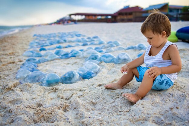 Ein Junge wird mit Muscheln und Sand gespielt, während er an einem mit Quallen übersäten Strand in der Nähe des Meeres sitzt