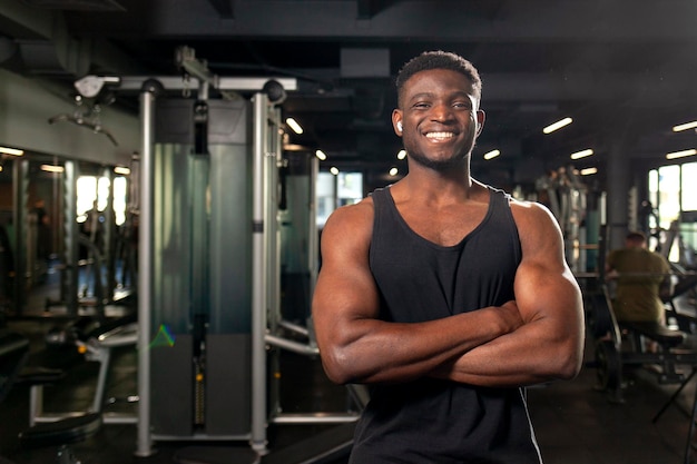 Ein junger afroamerikanischer Mann steht mit gekreuzten Armen in einem dunklen Fitnessstudio und lächelt dem Fitnesstrainer