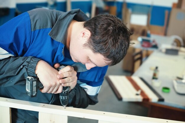 Ein junger Arbeiter in Overalls mit einem Schraubendreher sammelt Möbel in einer Tischlerei Hintergrund für die Herstellung von Möbeln