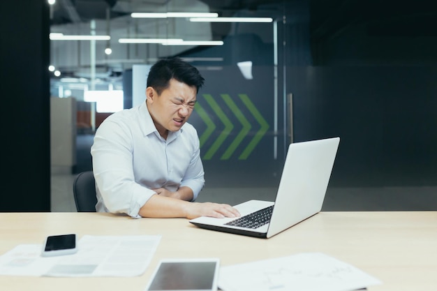 Ein junger asiatischer geschäftsmann hält sich am schreibtisch im büro den bauch und fühlt sich schlecht, fühlt schmerzen