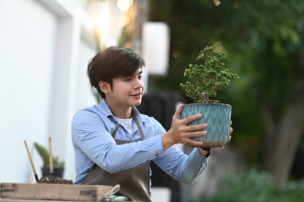 Ein junger asiatischer Mann, der Bonsai-Baumtopf hält, während er an seinem Hausgarten sitzt.