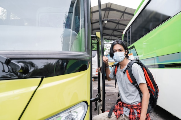 Ein junger asiatischer Mann mit Gesichtsmasken, der in den Bus steigt. Ein Mann, der eine Gesichtsmaske trägt und einen Rucksack trägt, steigt am Terminal in den Bus