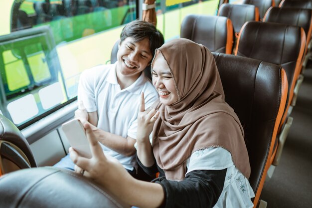 Ein junger asiatischer Mann und eine junge Frau in einem Hijab lächeln in die Kamera auf ihrem Handy, während sie zusammen ein Selfie machen, während sie im Bus sitzen