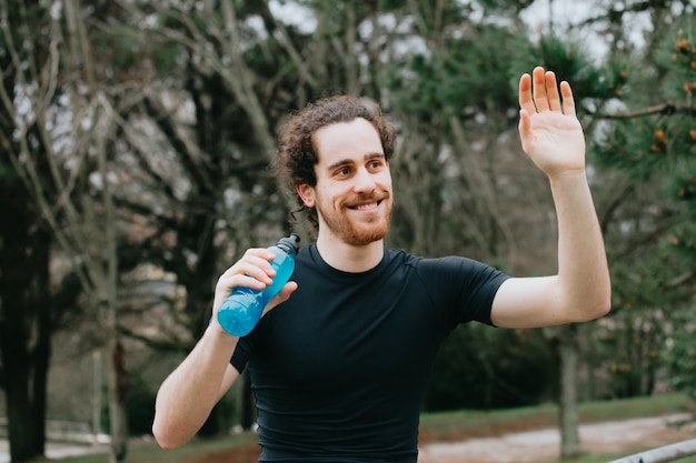 Ein junger athletischer Mann lächelt und grüßt, während er ein Karbonatgetränk im Park hält, während er Fitness macht