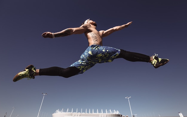 Ein junger athletischer Mann mit nacktem Oberkörper mit Stirnband in schwarzen Leggings und blauen Shorts springt an einem warmen sonnigen Tag hoch auf den Hintergrund des Stadions und des blauen Himmels.