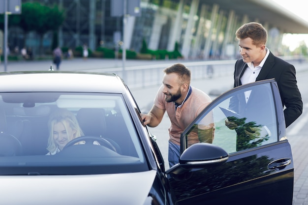 Foto ein junger autohändler in einem business-anzug zeigt den käufern ein neues auto. junges paar, mann und frau, kaufen ein auto. frau sitzt hinter dem lenkrad. kauf von maschinen, probefahrt.