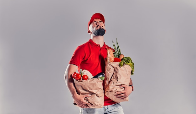 Ein junger bärtiger Kurier in rotem T-Shirt und Mütze hält zwei sehr schwere Lebensmittelsäcke. Essen nach Hause liefern.