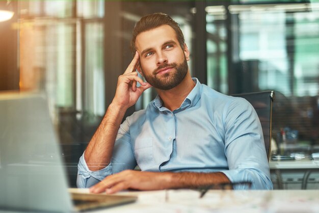 Ein junger bärtiger Mann in formeller Kleidung berührt seinen Kopf, lächelt und blickt weg, während er in einem modernen Büro arbeitet.