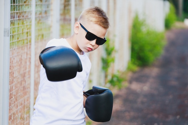 Ein junger blonder Junge in einem weißen T-Shirt und einer Sonnenbrille schlägt mit einer Hand in einem Boxhandschuh. Foto in hoher Qualität