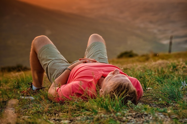 Ein junger blonder Mann liegt bei Sonnenuntergang auf dem Gras am Hang des Berggipfels