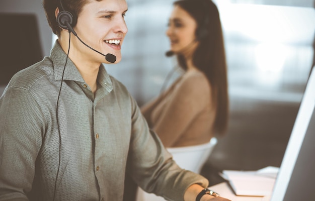 Ein junger dunkelhaariger Typ in einem grünen Hemd und Headsets spricht mit einem Kunden, während er am Schreibtisch sitzt und zusammen mit einer Kollegin in einem modernen Büro arbeitet.