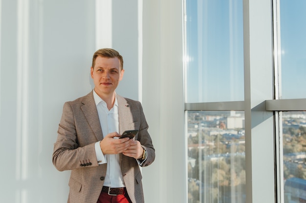 Ein junger erfolgreicher Geschäftsmann hält das Telefon in einem Büro mit Panoramafenstern in der Hand