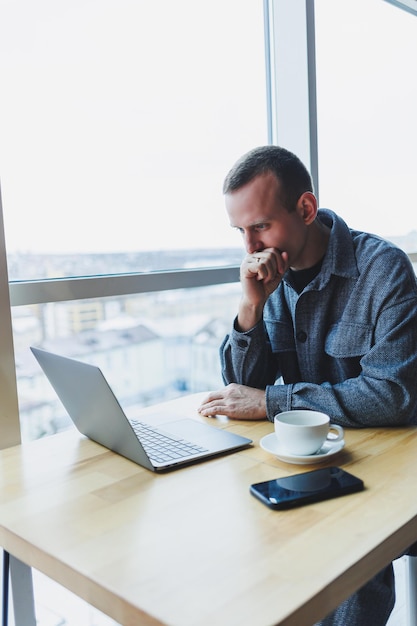 Ein junger erfolgreicher Geschäftsmann in einem Anzug trinkt Kaffee und arbeitet in einem Café im drahtlosen Internet auf einem Laptop, der allein an einem Tisch in einem Café sitzt