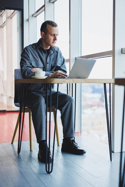 Ein junger erfolgreicher Geschäftsmann in einem Anzug trinkt Kaffee und arbeitet in einem Café im drahtlosen Internet auf einem Laptop, der allein an einem Tisch in einem Café sitzt