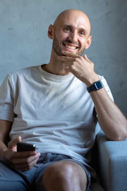 Ein junger, erfolgreicher, stilvoller Mann mit einem sauber rasierten Kopf Porträt eines Geschäftsmannes, der mit einem Smartphone in der Hand auf einem Stuhl sitzt