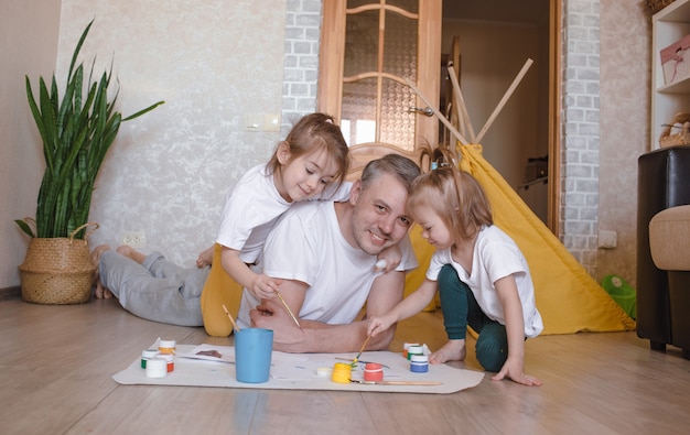 Ein junger fürsorglicher Vater lernt mit seinen jungen Töchtern malen, ein Mann liegt auf dem Boden und bringt Mädchen bei, Blumen und Einrichtungsgegenstände zu zeichnen.