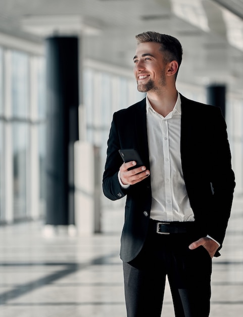 Ein junger Geschäftsmann in einem Business Center in schwarzem Anzug und weißem Hemd mit einem Telefon in der Hand und einer Hand in der Tasche lächelt und schaut weg