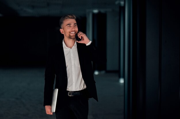 Ein junger Geschäftsmann in einem Business Center mit einem Telefon und einem Laptop in der Hand in einem schwarzen Anzug und weißem Hemd lächelt und spricht am Telefon