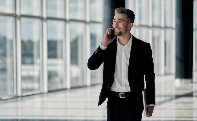 Ein junger Geschäftsmann in einem Business Center mit einem Telefon und einem Laptop in der Hand in einem schwarzen Anzug und weißem Hemd lächelt und spricht am Telefon