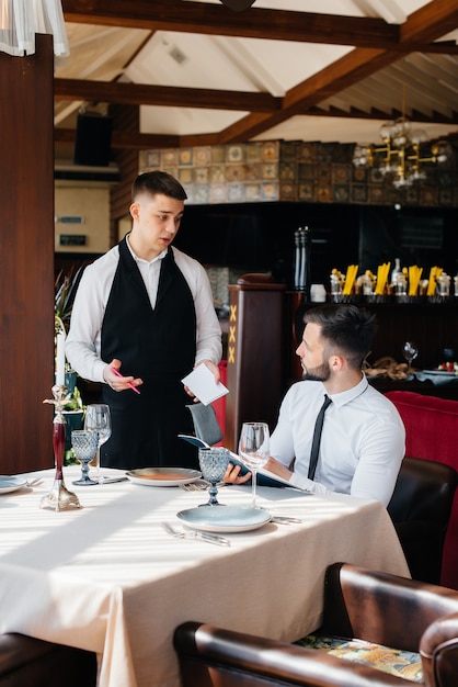 Ein junger Geschäftsmann in einem feinen Restaurant begutachtet die Speisekarte und bestellt einem jungen Kellner in stilvoller Schürze. Kundendienst.