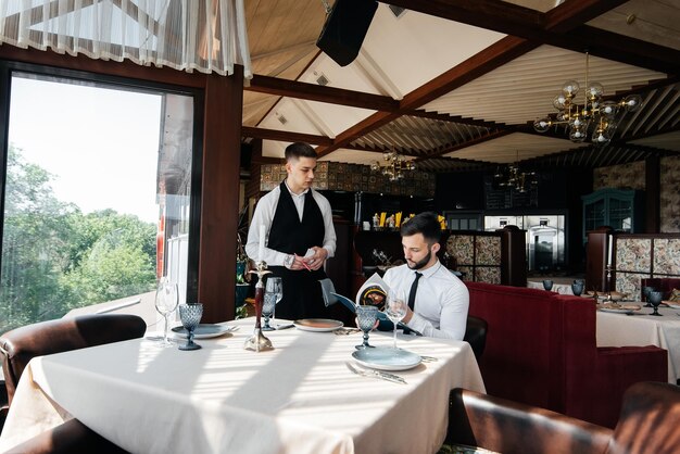 Ein junger Geschäftsmann in einem feinen Restaurant prüft die Speisekarte und bestellt bei einem jungen Kellner in stilvoller Schürze. Kundenservice Tischservice im Restaurant