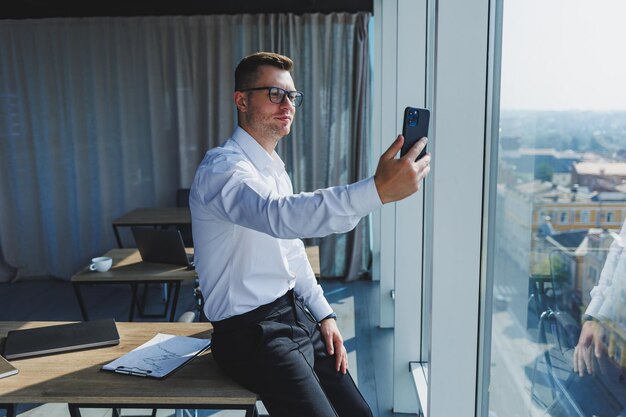 Ein junger Geschäftsmann mit einem Telefon in seinen Händen in Gläsern und einem weißen Hemd ist er in einem modernen Büro mit großen Fenstern Ein Mann in klassischen Hosen und einem weißen Hemd Junger attraktiver Geschäftsmann