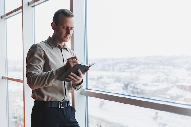Ein junger Geschäftsmann steht an einem Panoramafenster in einem Wolkenkratzer und macht sich mit einem Stift Notizen in seinem Notizbuch Ein Mann in Hemd und Hose mit europäischem Aussehen