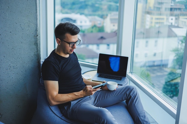 Ein junger Geschäftsmann sucht nach etwas und tippt auf einem Smartphone und trinkt frischen schwarzen Kaffee Das Konzept einer modernen erfolgreichen Person Junger konzentrierter Mann mit Brille in einem offenen Büro