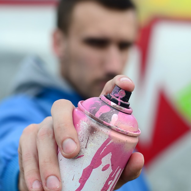 Ein junger Graffitikünstler in einem Matrosenmantel hält eine Dose Farbe vor ihm vor einem Hintergrund der farbigen Graffitizeichnung. Straßenkunst- und Vandalismuskonzept