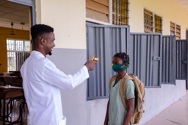 Ein junger gutaussehender afrikanischer klassenlehrer, der als thermometer hält, um die temperatur seines schülers zu scannen, bevor er das klassenzimmer betritt