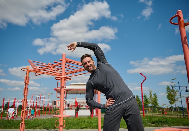 Ein junger gutaussehender europäischer Kaukasier von athletischer Statur treibt Sport auf einem Sommersportplatz, führt Seitenbeugen aus und streckt seinen Arm aus.