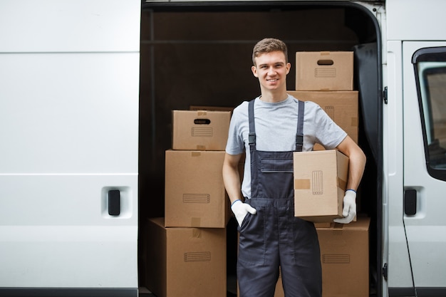 Ein junger, gutaussehender, lächelnder Arbeiter in Uniform steht neben dem Lieferwagen voller Kisten, der eine Kiste in den Händen hält. Umzug, Umzugsservice.