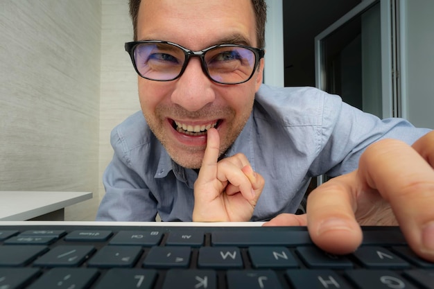 Ein junger gutaussehender Mann in einem blauen Hemd arbeitet vor einem hellen Hintergrund an einem Computer. Der Nerd blickt auf den Bildschirm und stochert mit dem Finger in den Zähnen