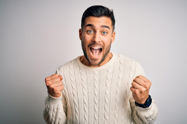 Ein junger, gutaussehender Mann in einem gewöhnlichen Pullover steht über einem isolierten weißen Hintergrund und feiert überrascht und erstaunt über den Erfolg mit erhobenen Armen und offenen Augen.