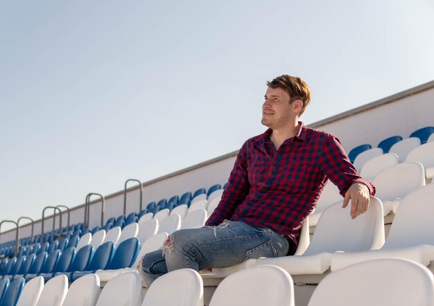 Ein junger gutaussehender mann in kariertem hemd und jeans sitzt allein auf einer stadiontribüne, lächelt und schaut im sommer weg