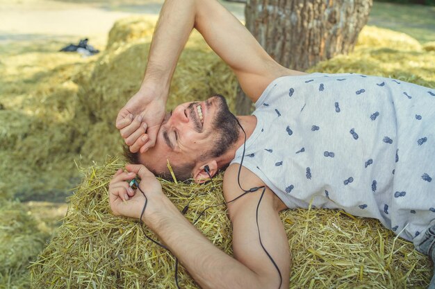 Foto ein junger gutaussehender mann mit bart in t-shirt und shorts liegt lächelnd im heu und hört mit geschlossenen augen musik