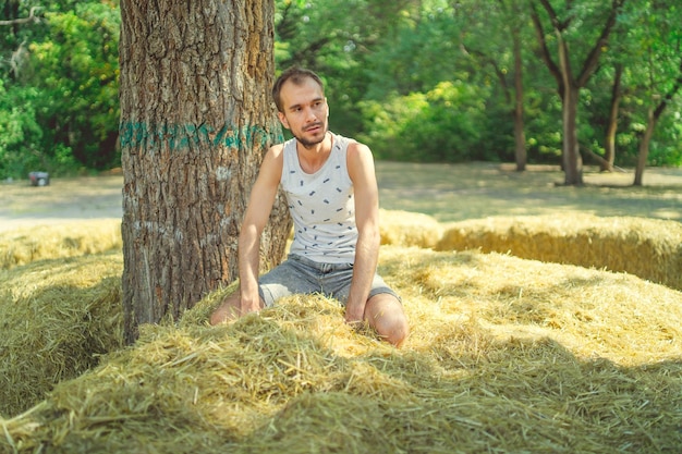 Ein junger gutaussehender Mann mit schlankem Bart sitzt im Heu und hält Heu in den Händen vor dem Hintergrund grüner Bäume im Park