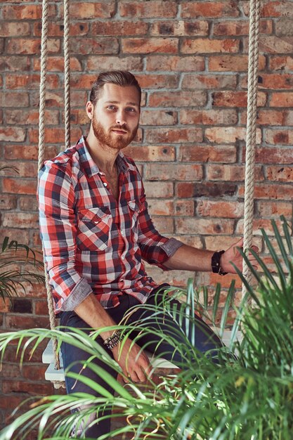 Ein junger gutaussehender rothaariger Modellmann sitzt auf einer Schaukel im Raum mit Loft-Interieur.