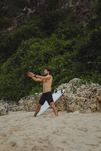 Ein junger gutaussehender Surfer am Meeresufer wärmt sich vor dem Surfen auf. Übungen vor dem Sport, Stretching vor dem Surfen.