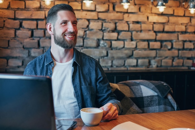 Ein junger Hipster in einer Bar trinkt einen Cappuccino. Ein junger Mann trinkt Kaffee in einem Stadtcafé während der Mittagspause und arbeitet an einem Laptop