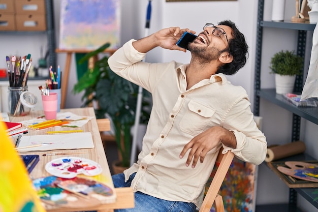 Ein junger hispanischer Künstler lächelt zuversichtlich und spricht im Kunststudio mit dem Smartphone