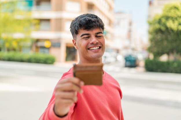 Foto ein junger hispanischer mann hält eine brieftasche im freien mit einem glücklichen gesichtsausdruck