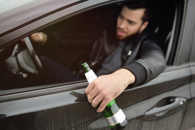 Ein junger hübscher bärtiger Mann mit einer Flasche Bier oder einem alkoholarmen Getränk am Steuer eines Autos. Fahren in einem Zustand der Vergiftung