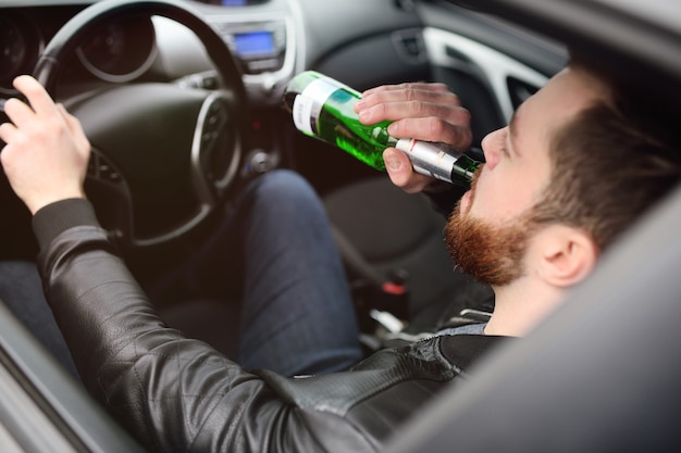 Ein junger hübscher bärtiger Mann mit einer Flasche Bier oder einem alkoholarmen Getränk am Steuer eines Autos. Fahren in einem Zustand der Vergiftung