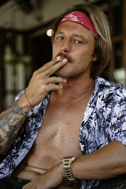 Ein junger hübscher Hippie-Mann in einem roten Kopftuch mit nacktem Oberkörper rollt in einem Café in Bali eine Tabakzigarette.