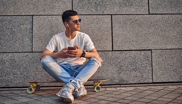 Ein junger kaukasischer Amerikaner mit Brille sitzt im Schneidersitz auf einem Longboard und hält ein Telefon