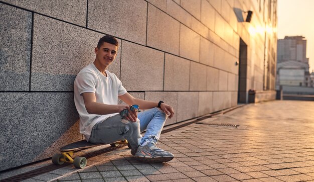 Ein junger kaukasischer Amerikaner sitzt im Schneidersitz auf einer Longboard-Brille in der Hand an der Wand