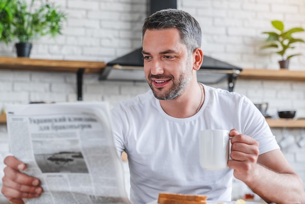 Ein junger kaukasischer Mann liest Zeitung, während er in einer modernen Küche frühstückt